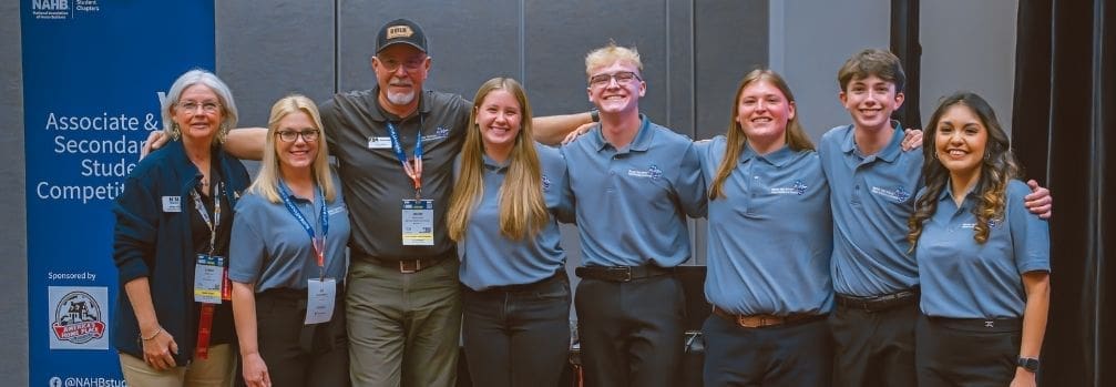 Group of students and mentors standing together and smiling at the NAHB Associate and Secondary Student Competition event, wearing matching blue shirts.