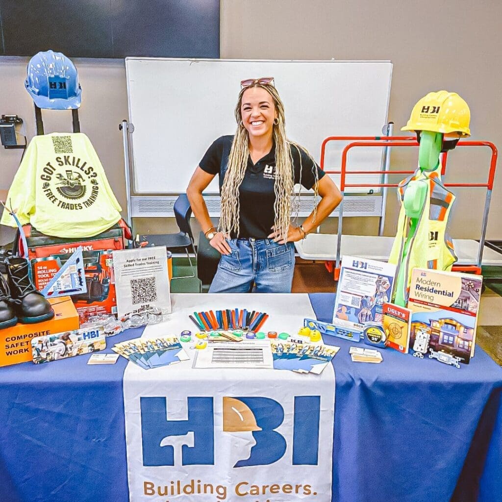 Smiling woman standing behind an HBI Building Careers booth with training materials, safety gear, hard hats, books, and promotional items displayed on the table.